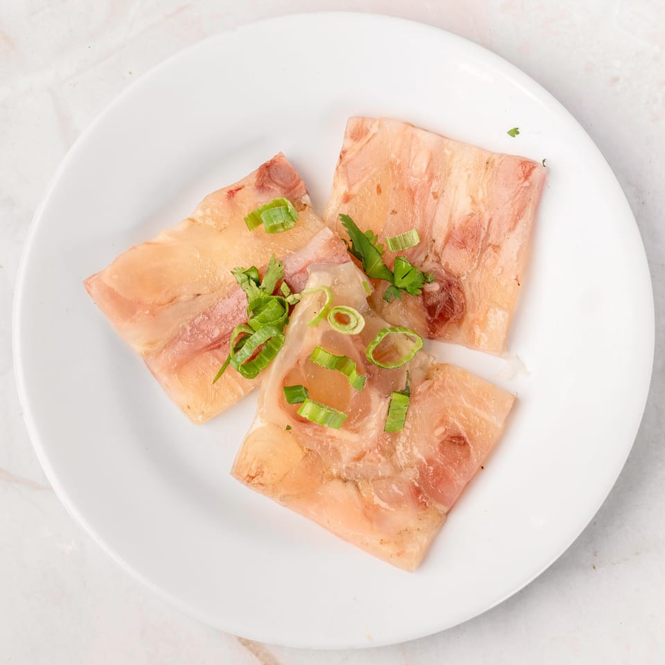 Three slices of translucent meat jelly, garnished with fresh green onions and cilantro, on a white plate.