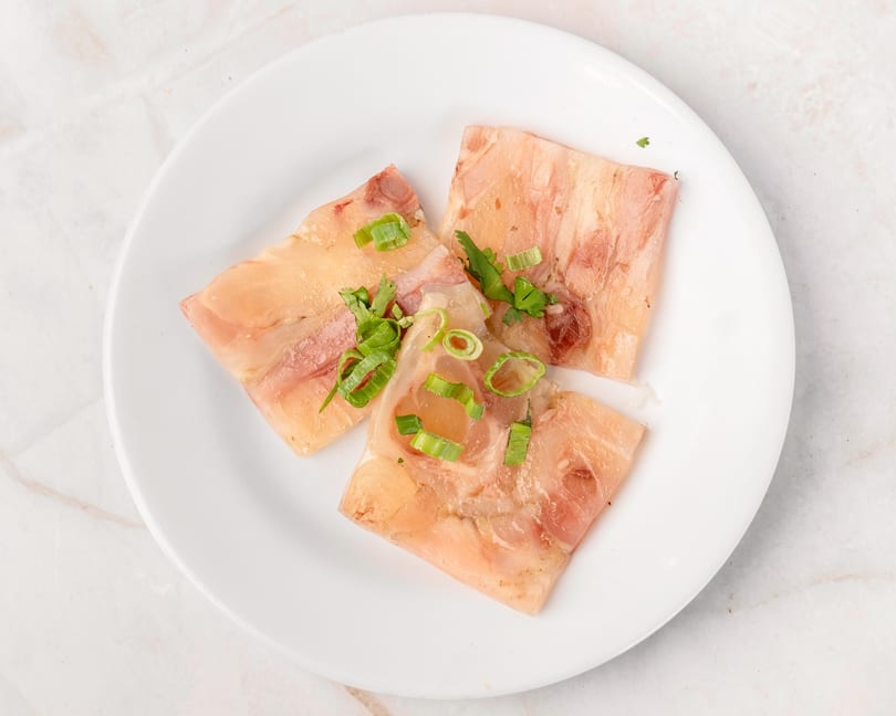 Three slices of translucent meat jelly, garnished with fresh green onions and cilantro, on a white plate.