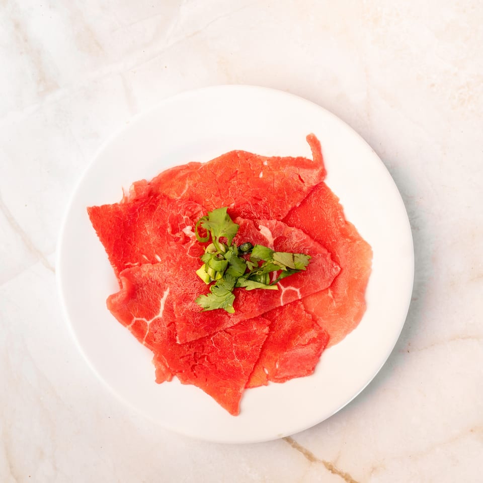 Thinly sliced raw beef, garnished with fresh green herbs, on a white plate.
