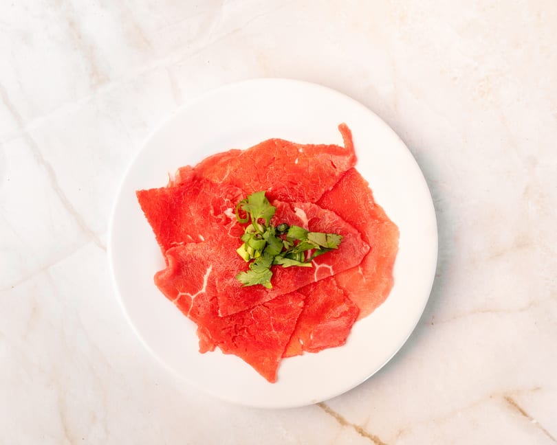 Thinly sliced raw beef, garnished with fresh green herbs, on a white plate.