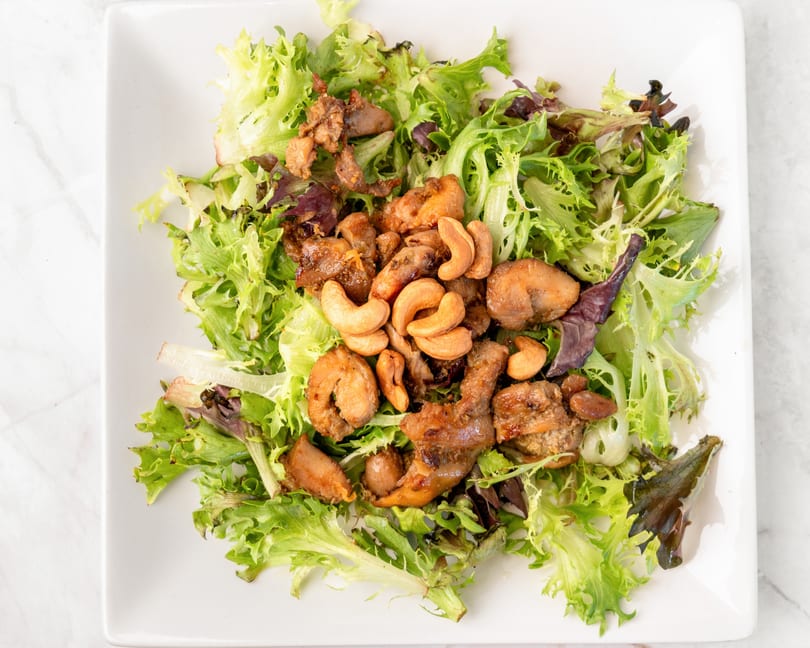 A white square plate holds a fresh green salad topped with chicken and cashews.