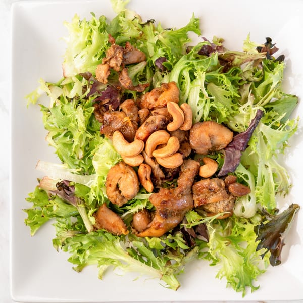 A square white plate filled with green and purple lettuce, topped with chicken and cashews.