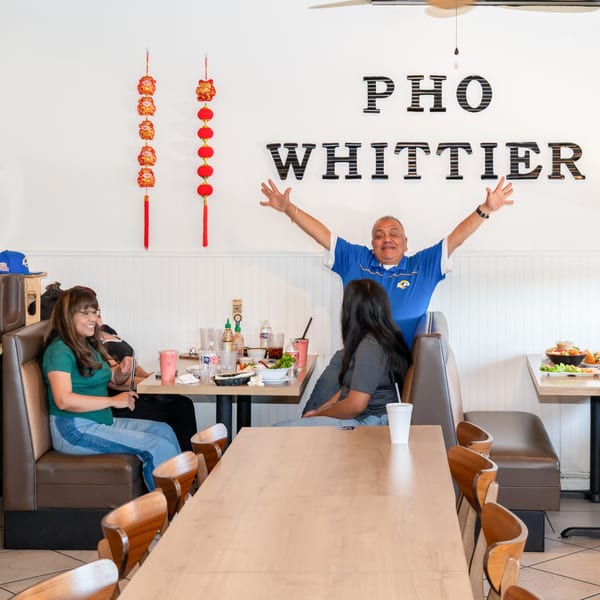 A man with arms outstretched behind two women dining at Pho Whittier, with food and drinks on tables.