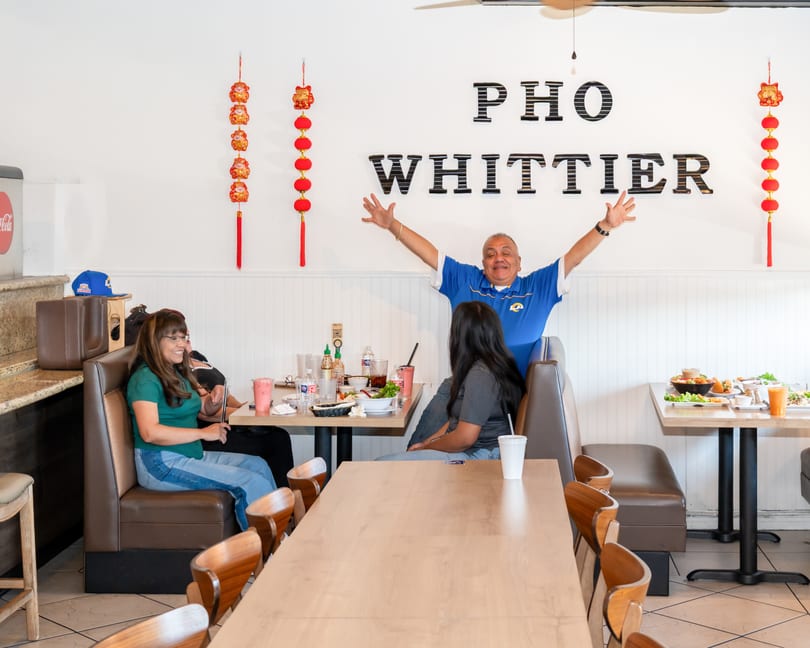 A man with arms outstretched behind two women dining at Pho Whittier, with food and drinks on tables.
