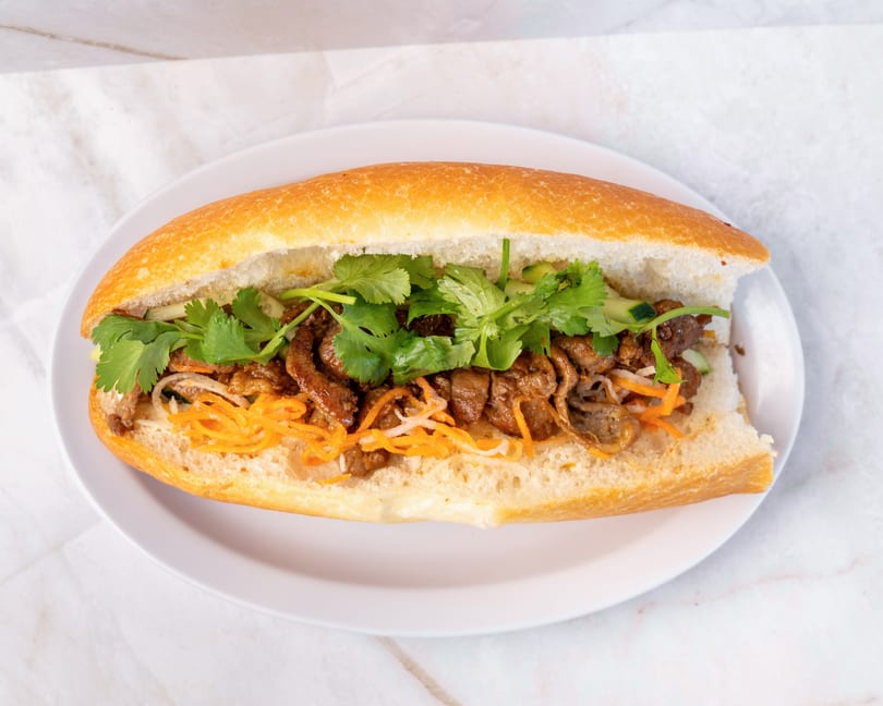 A Vietnamese banh mi sandwich with grilled meat, pickled carrots, and fresh cilantro on a white plate.