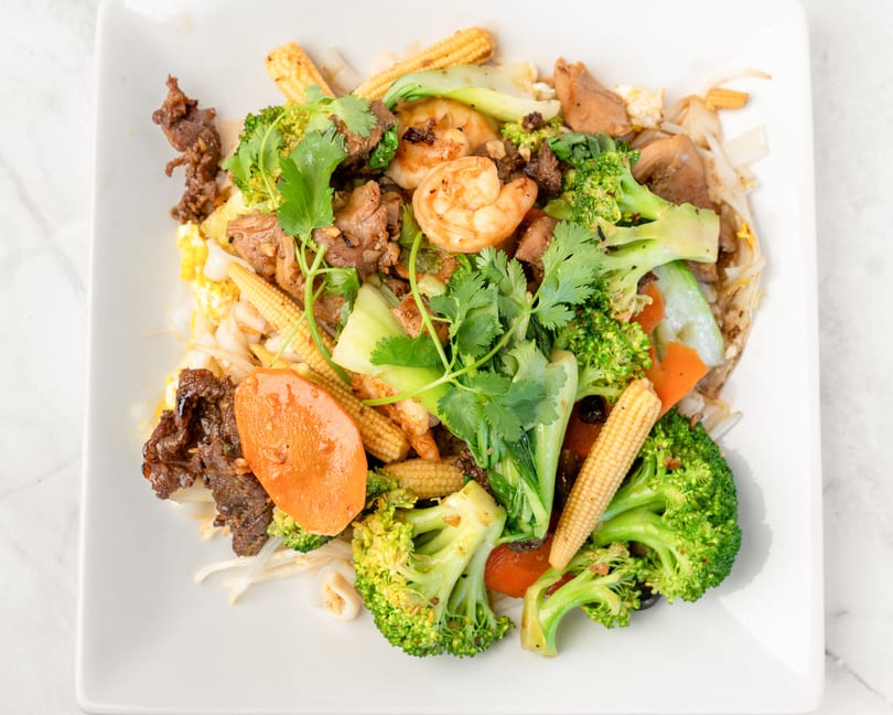 Stir-fried noodles with beef, shrimp, broccoli, baby corn, and carrots on a white plate.