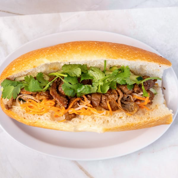 A Vietnamese banh mi sandwich filled with grilled meat, pickled carrots, and fresh cilantro on a white plate.