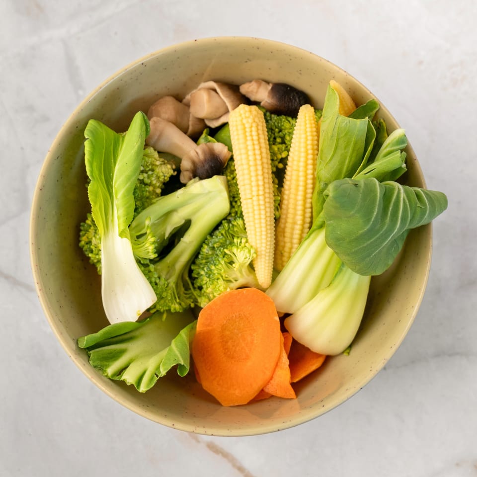 A bowl of fresh vegetables including broccoli, baby corn, bok choy, carrots, and mushrooms.