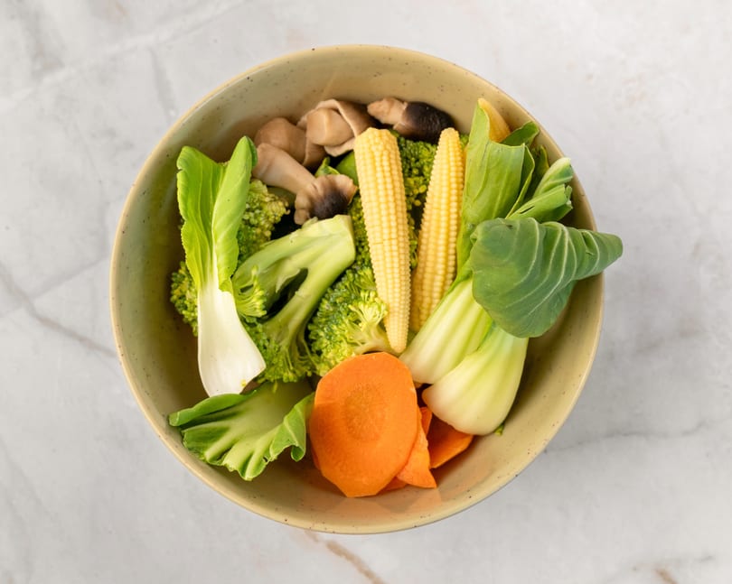 A bowl of fresh vegetables including broccoli, baby corn, bok choy, carrots, and mushrooms.