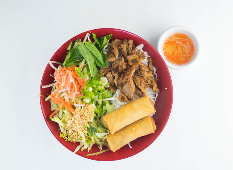 Vietnamese vermicelli bowl with grilled pork, spring rolls, fresh vegetables, and a side of sauce.