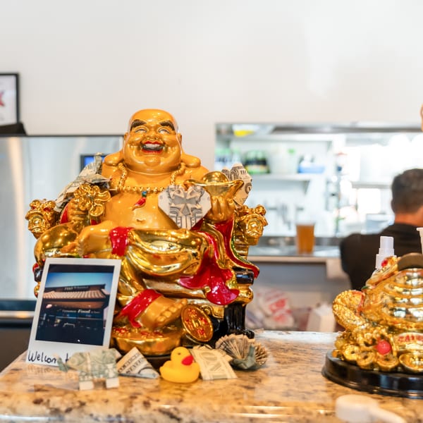 A golden Buddha statue with money and a polaroid on a counter, with a kitchen in the background.
