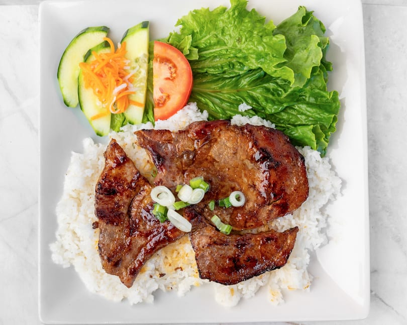 Grilled pork chops with rice, lettuce, tomato, cucumber, and pickled carrots on a white plate.