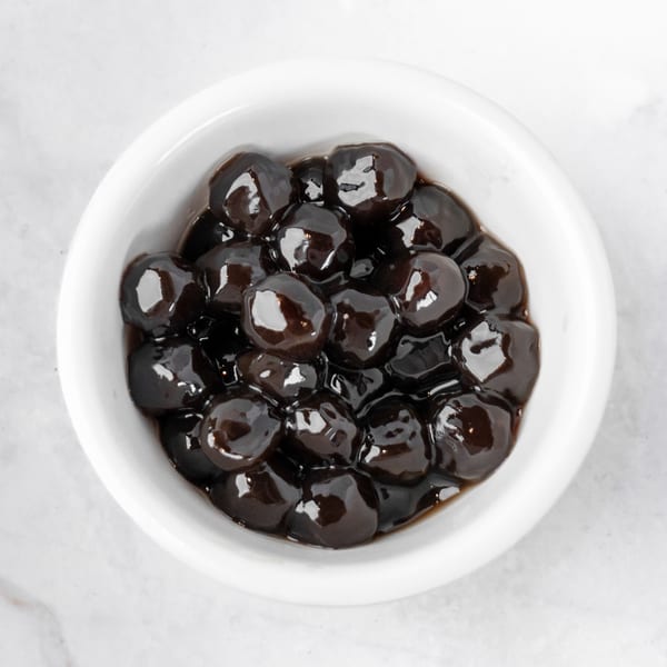 A white bowl filled with dark, glossy tapioca pearls, also known as boba, on a white marble surface.