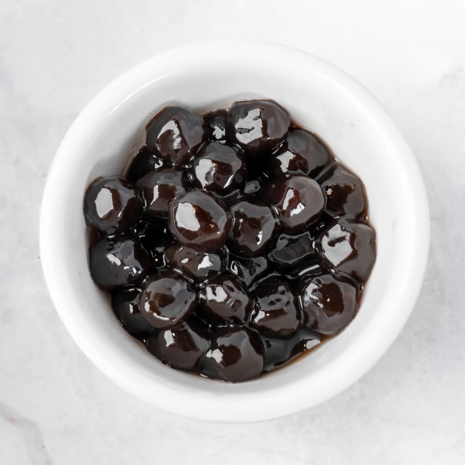 A white bowl filled with dark, glossy tapioca pearls, also known as boba, on a white marble surface.