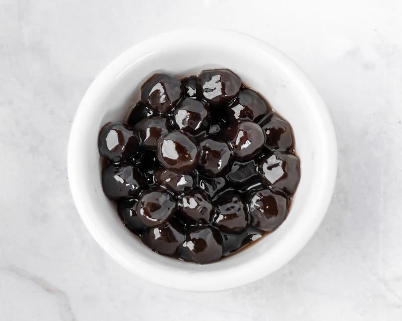 A white bowl filled with dark, glossy tapioca pearls, also known as boba, on a white marble surface.