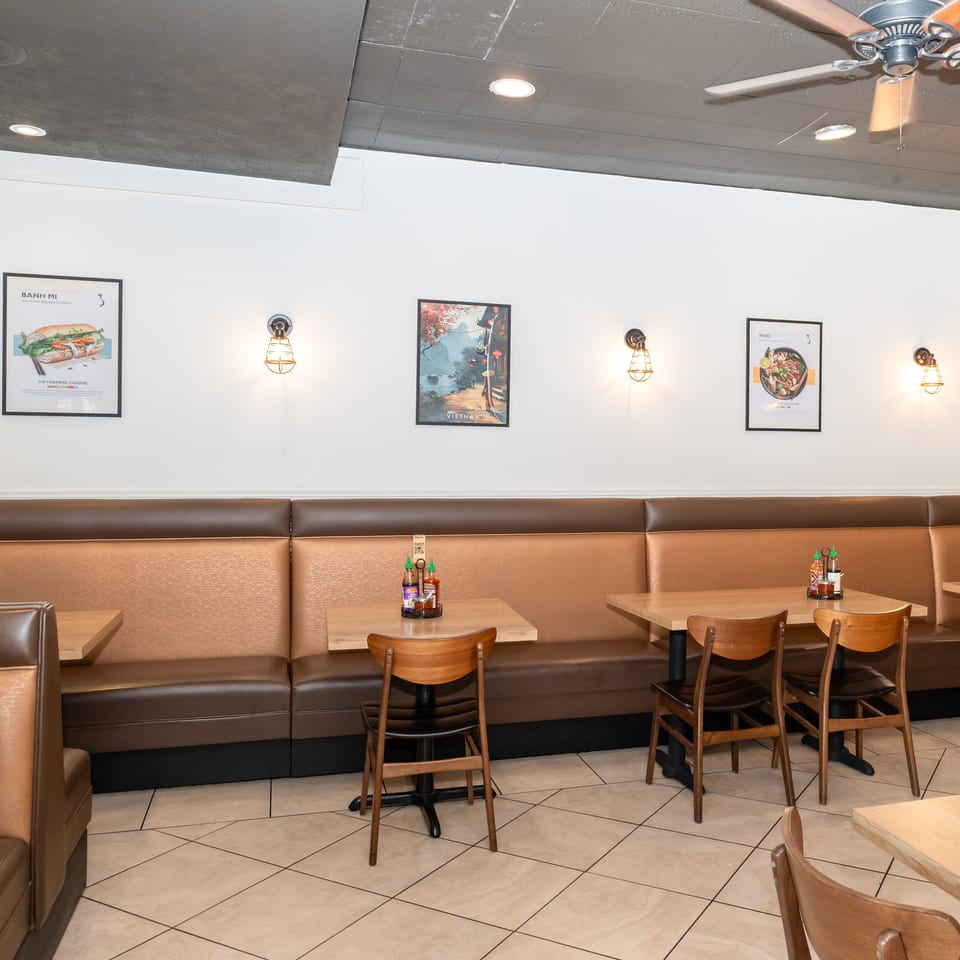 Interior of a Vietnamese restaurant with brown booths, light wooden tables, and food posters on white walls.