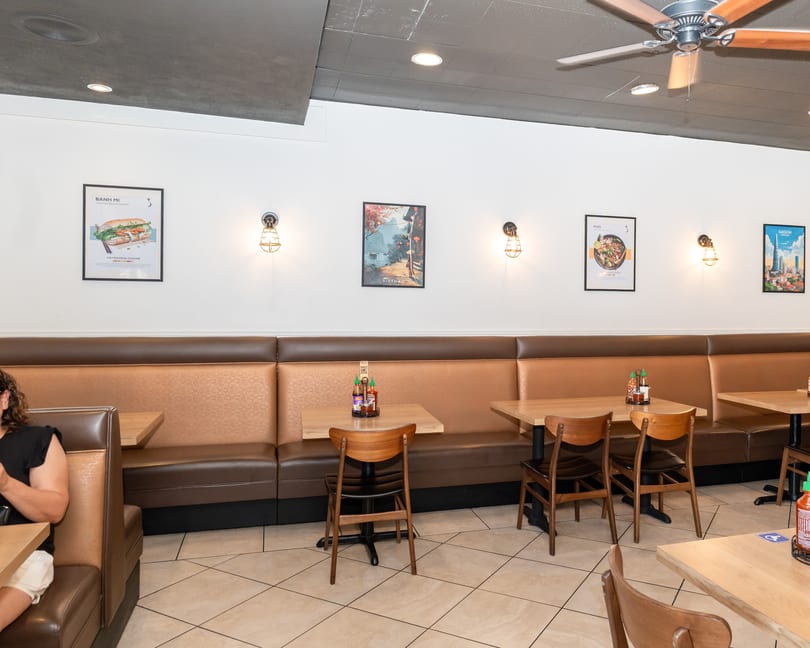 Interior of a Vietnamese restaurant with brown booths, light wooden tables, and food posters on white walls.