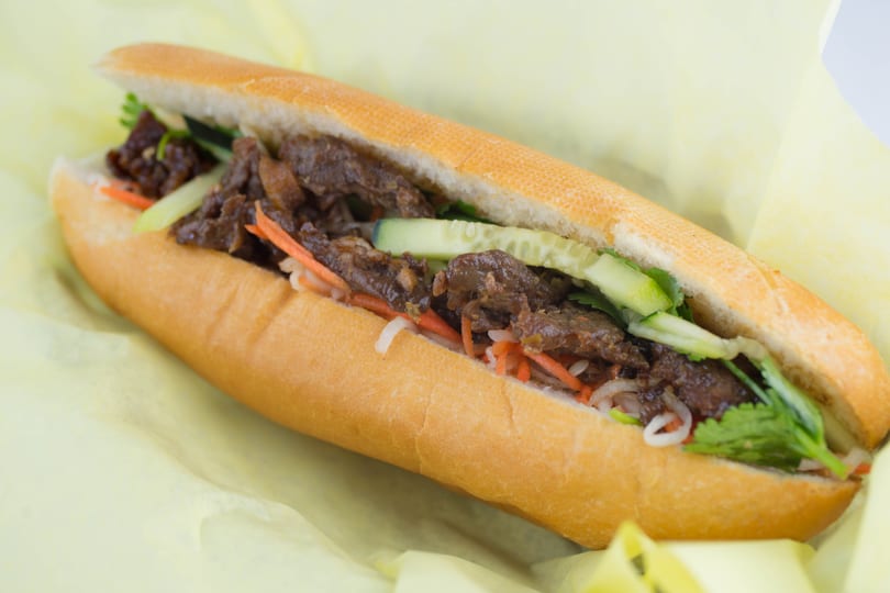 A banh mi sandwich filled with beef, pickled carrots, cucumbers, and cilantro on yellow paper.