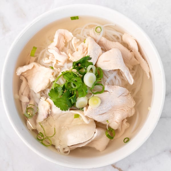 A white bowl of chicken pho with noodles, broth, cilantro, and green onions on a marble surface.