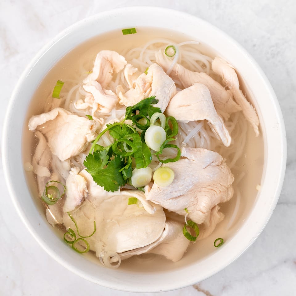 A white bowl of chicken pho with noodles, broth, cilantro, and green onions on a marble surface.