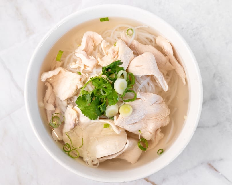 A white bowl of chicken pho with noodles, broth, cilantro, and green onions on a marble surface.